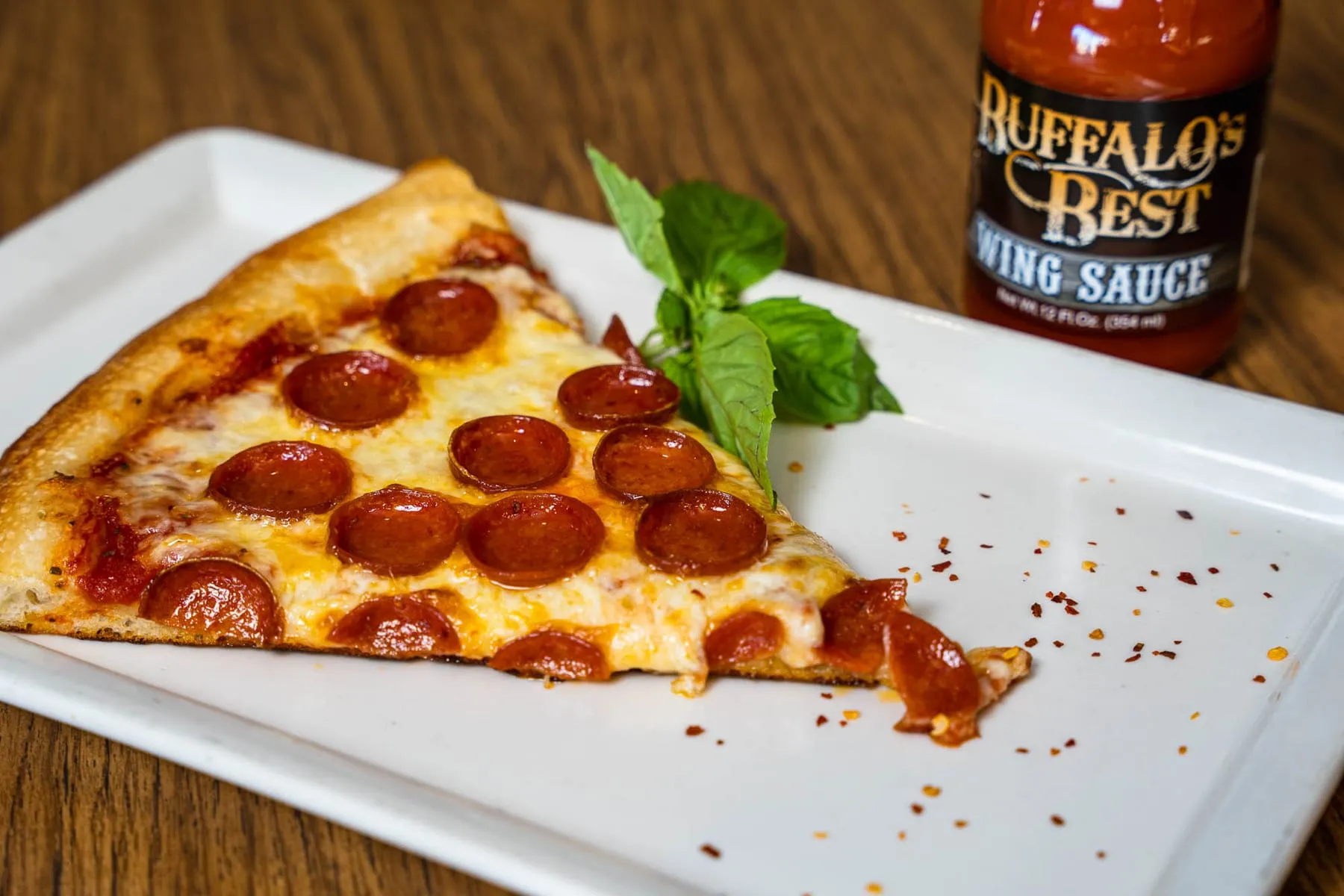 A slice of pepperoni pizza with a bottle of wing sauce